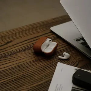 KeyBudz Artisan Series brunt læderetui til AirPods Pro åbent på et skrivebord med øretelefoner og en bærbar computer i nærheden