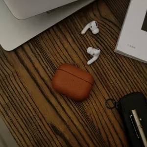 KeyBudz brunt læderetui til AirPods Pro på et skrivebord med øretelefoner, med et hængslet design med hård skal.