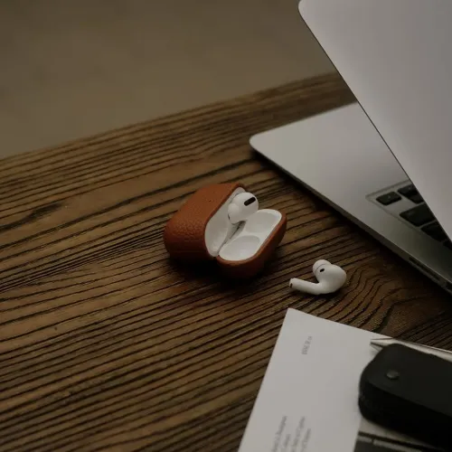 Nahast ümbris AirPods Pro Gen 2 jaoks naturaalse pruuni värvi, avatud puidust laual; valge laadimiskork nähtav ja pistik on laual.