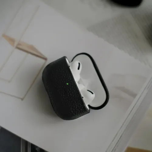 Svart räfflat läderfodral för AirPods Pro med öppet lock och synlig grön indikator, placerat på en tjock bok i mjukt ljus.