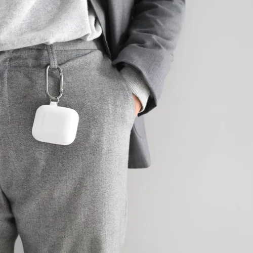 Vitt AirPods-skal i silikon med karbinhake, fäst i byxhälla; fyrkantig form med lock och matt yta, hängande mot grå byxa.