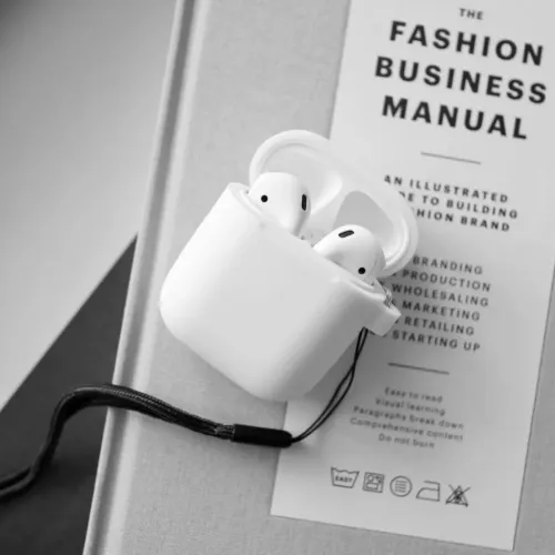 Vitt silikonskal för AirPods med öppet lock och svart handledsrem fäst i hörnet, avbildat liggande på en bok i närbild.