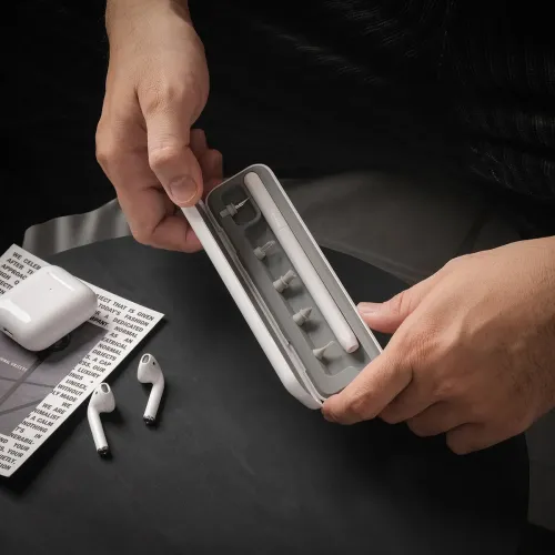 AirCare 2.0-rengøringssæt i hvidt etui åbnes; penlignende værktøj og udskiftelige spidser er synlige, med AirPods og etui på bordet.