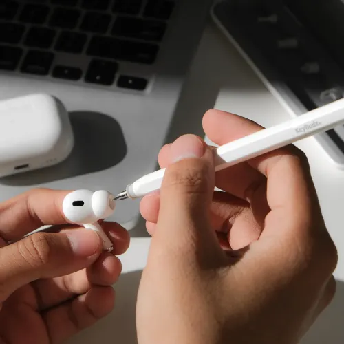 KeyBudz AirCare 2.0 rengøringsværktøj med spids bruges til at rengøre en hvid øreprop; etui og bærbar computer i baggrunden.