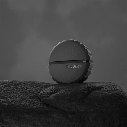 KeyBudz Covert Mount for AirTag, svart, rund holder med rillet kant, frontspor og gravert logo, står på en stein.