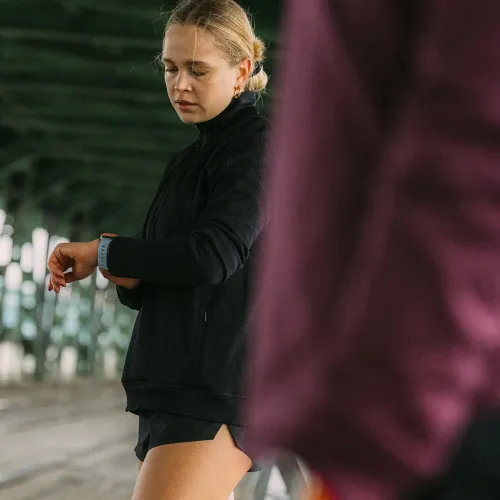 Apple Watch-bånd i blått med perforert mønster, montert på Apple Watch på håndleddet; i bakgrunnen er det grønne bjelker i hallen