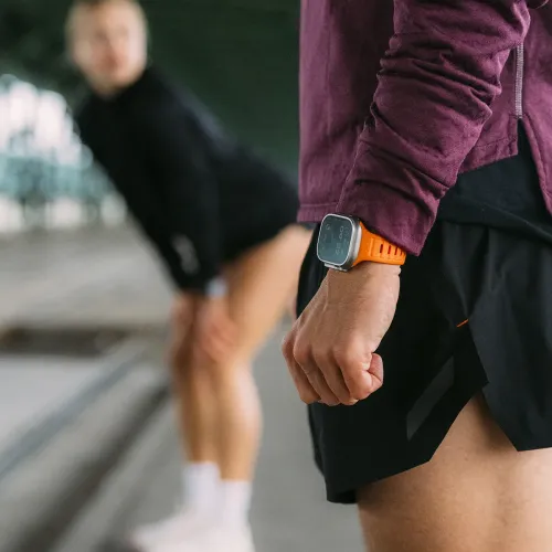 Apple Watch-bånd i elektrisk oransje med rektangulære spor, vist på et håndledd, med uskarp utendørs sporty bakgrunn.