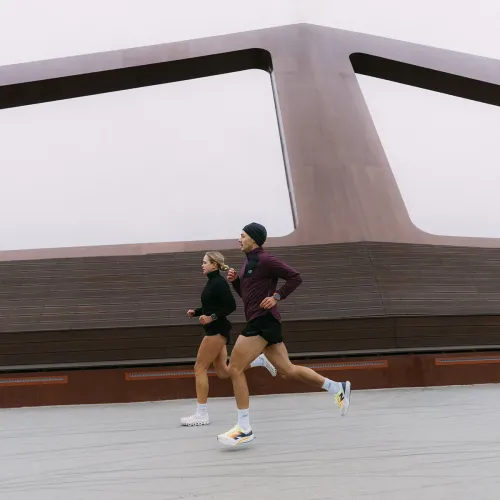 Elektrisk oransje Apple Watch-bånd synlig på venstre håndledd til en løper i profil, utendørs i bevegelse foran en rusten bygning.