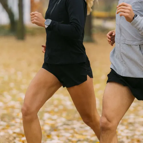 Vasemmassa ranteessa oleva kiviharmaa Apple Watch -ranneke, joka näkyy lähikuvassa puistoympäristössä hölkkäävästä mustiin vaatteisiin pukeutuneesta juoksijasta.