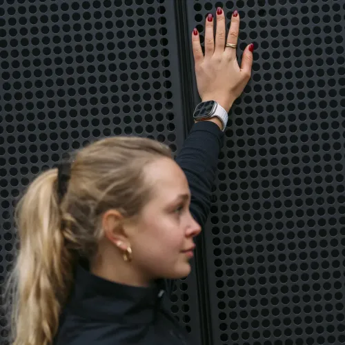 KeyBudz Journey Apple Watch -ranneke, kiviharmaa, sileä hihna ranteessa Apple Watchin kanssa, kuvassa rei'itettyä tummaa metalliseinää vasten.