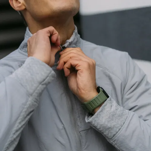 Apple Watch-armband i skogsgrönt med räfflat band på svart klocka syns på handled när person drar upp grå jackas dragkedja.